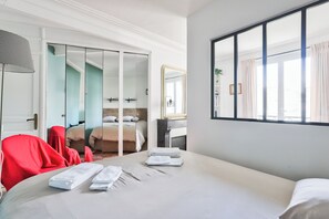 Ein Hotelzimmer mit Bett, einem roten Sessel, einem Spiegel und Blick auf eine Stadtlandschaft durch das Fenster.