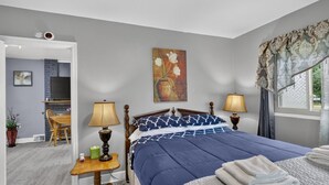 A bedroom with a bed, two bedside lamps, a painting, a window with curtains, and a small table with a lamp and a plant. - Cozy Private Home w Fire Pit Near Downtown! (Kalamazoo)