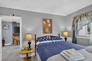 A bedroom with a bed, two bedside lamps, a painting, a window with curtains, and a small table with a lamp and a plant. - Cozy Private Home w Fire Pit Near Downtown! (Kalamazoo)
