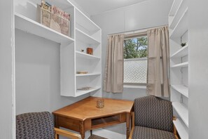 Una habitación de esquina con una mesa de madera y dos sillas, estanterías blancas y una ventana con cortinas.