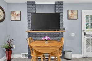 A dining area with a brick fireplace, a flat-screen TV mounted above a wooden table, and two chairs. - Cozy Private Home w Fire Pit Near Downtown! (Kalamazoo)