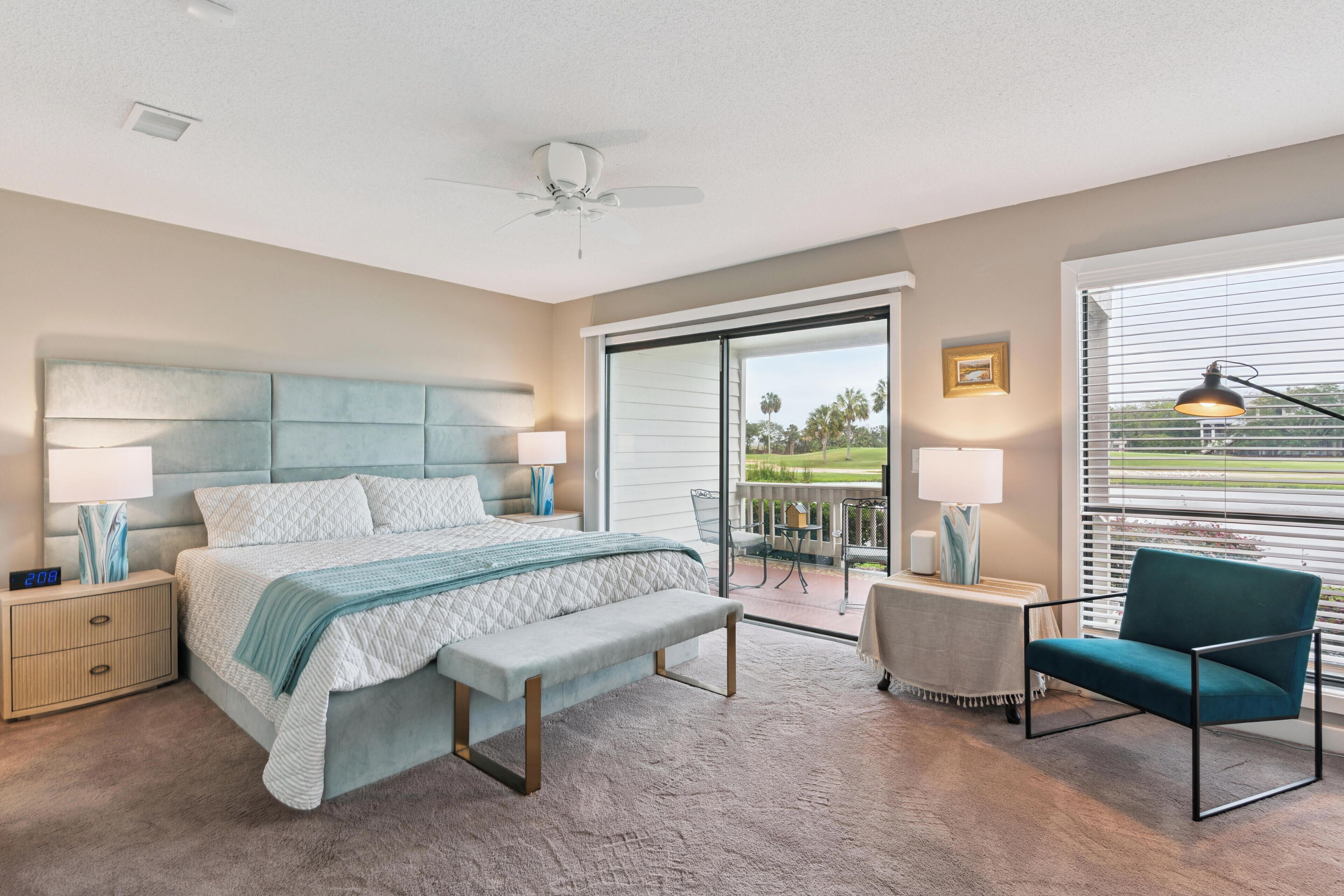 A bedroom with a bed, bedside tables, a bench, a chair, a ceiling fan, and a view through a sliding glass door.