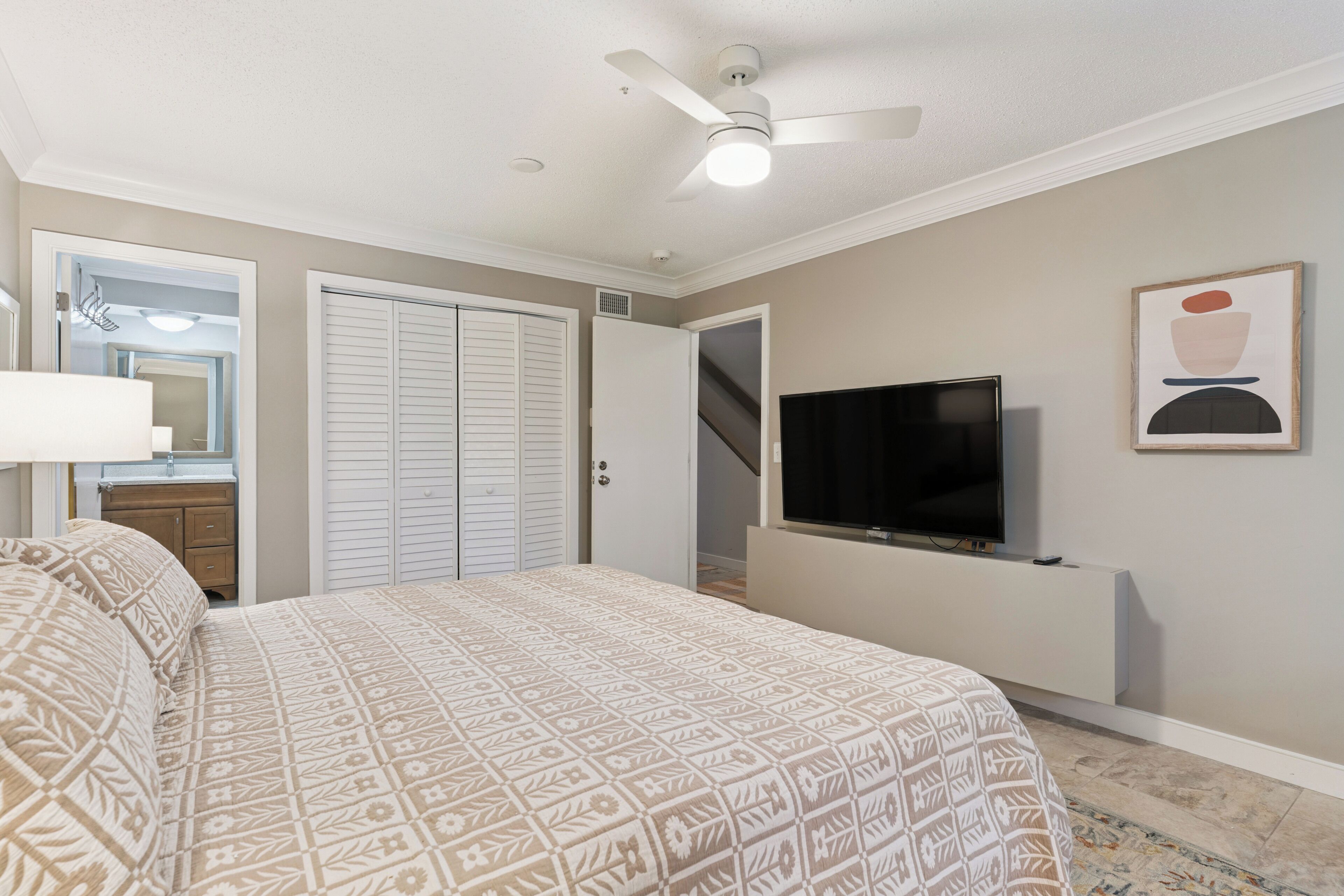Une chambre à coucher comprenant un lit, un ventilateur de plafond, un téléviseur fixé au mur, une porte et un miroir.