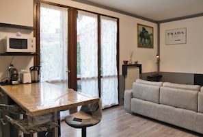 A kitchen with a microwave, toaster, and coffee maker, a dining table with a chair, a sofa, and a window with curtains. - Il Giardino sul Lago – Pool Residence (Garlate)