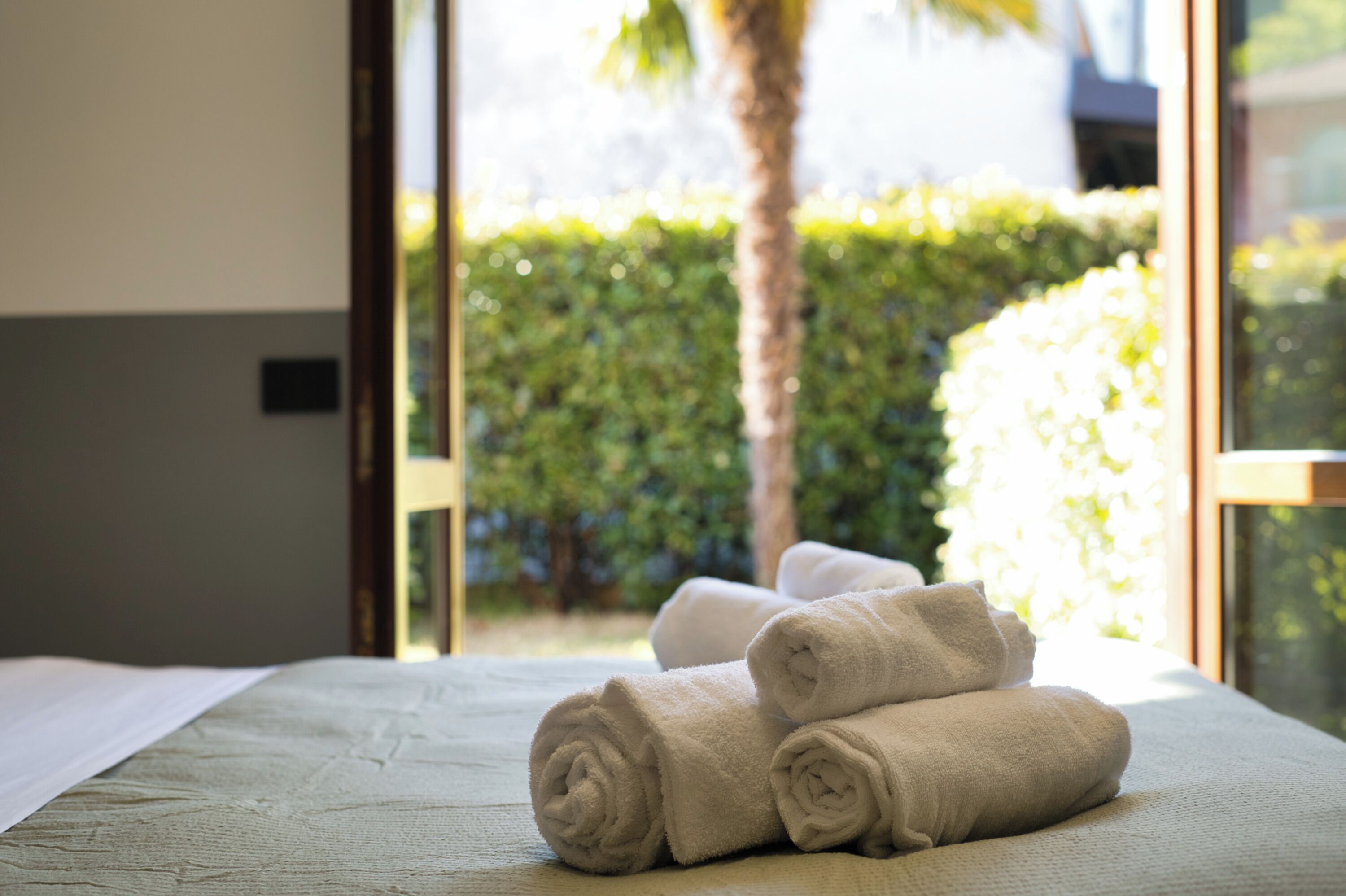A hotel room with a bed, a window with a view of greenery, and rolled towels on the bed.