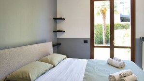 Une chambre à coucher avec un lit, des oreillers, une fenêtre donnant sur des palmiers et une porte.