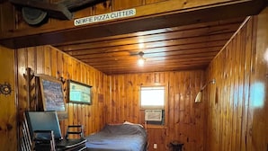 A wooden cabin room with a bed, a desk, a chair, a window, and a sign that reads 'NEWFIE PIZZA CUTTER'. - Rustic Fishing Lodge located on the Main Southwest Miramichi (Bloomfield Ridge)