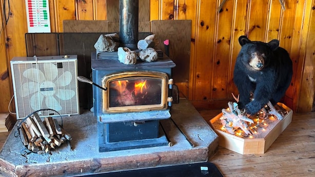 A wood-burning stove with logs, a bear figurine, and a thermostat on a wooden wall. - Rustic Fishing Lodge located on the Main Southwest Miramichi (Bloomfield Ridge)