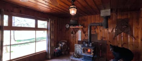 A wooden cabin interior with a fireplace, a ceiling fan, and a large window.