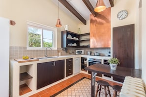 A modern kitchen with dark cabinetry, a window, a sink, a microwave, and a dining area with stools.