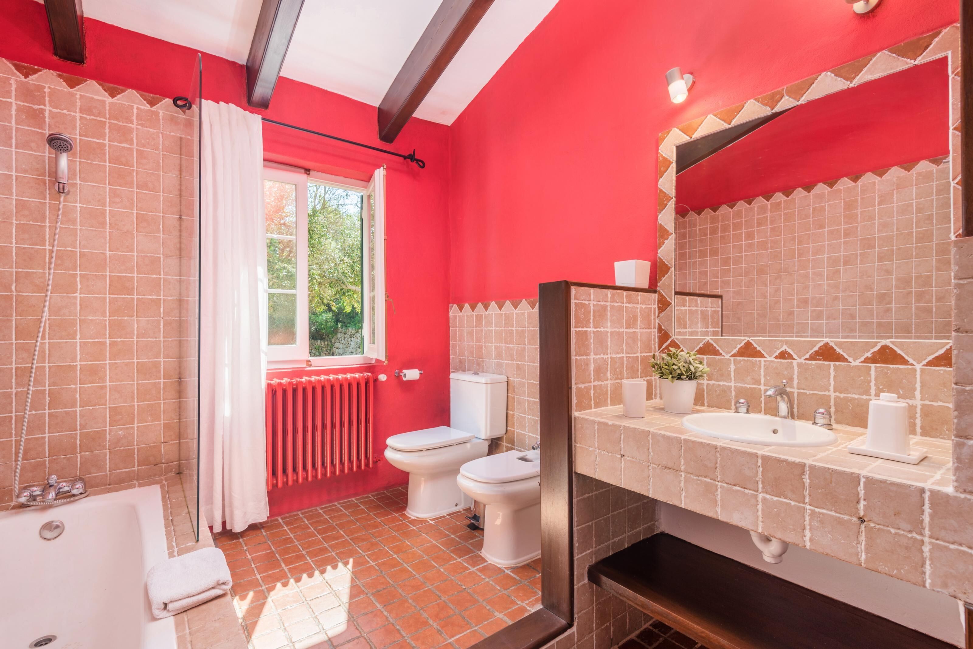 A bathroom with a red wall, a bathtub, two toilets, a sink, and a window with a view of greenery.
