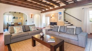 A living room with two sofas, a coffee table, wooden beams on the ceiling, a staircase, and a dining area in the background. - Villa Mig Coster - A rural retreat on a private estate (Sant Lluís)