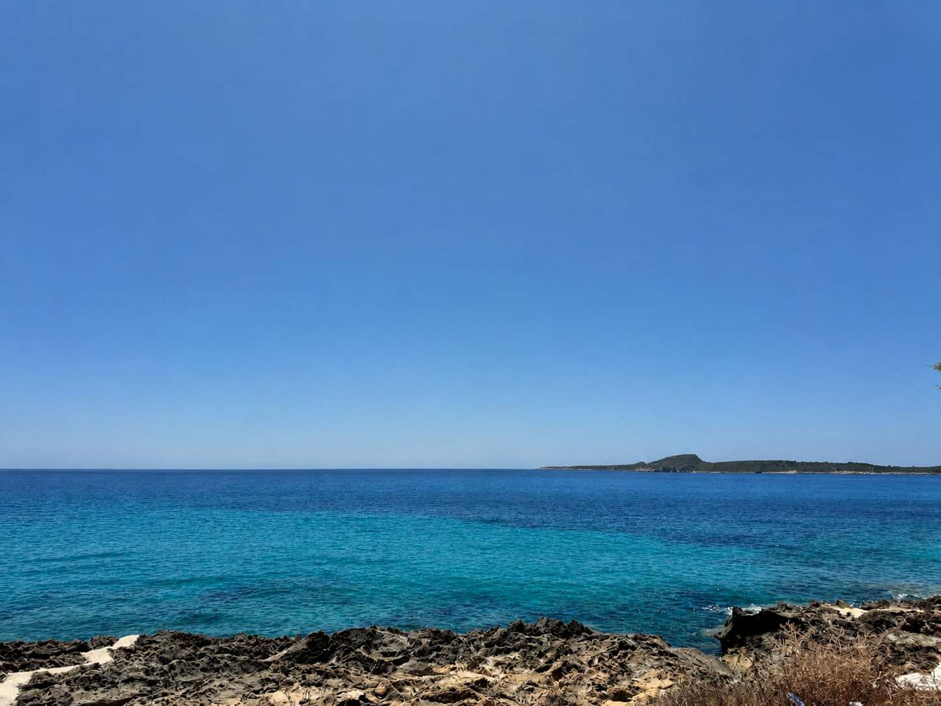 Paisagem costeira com águas azul-claras, terreno rochoso e formações terrestres ao longe.