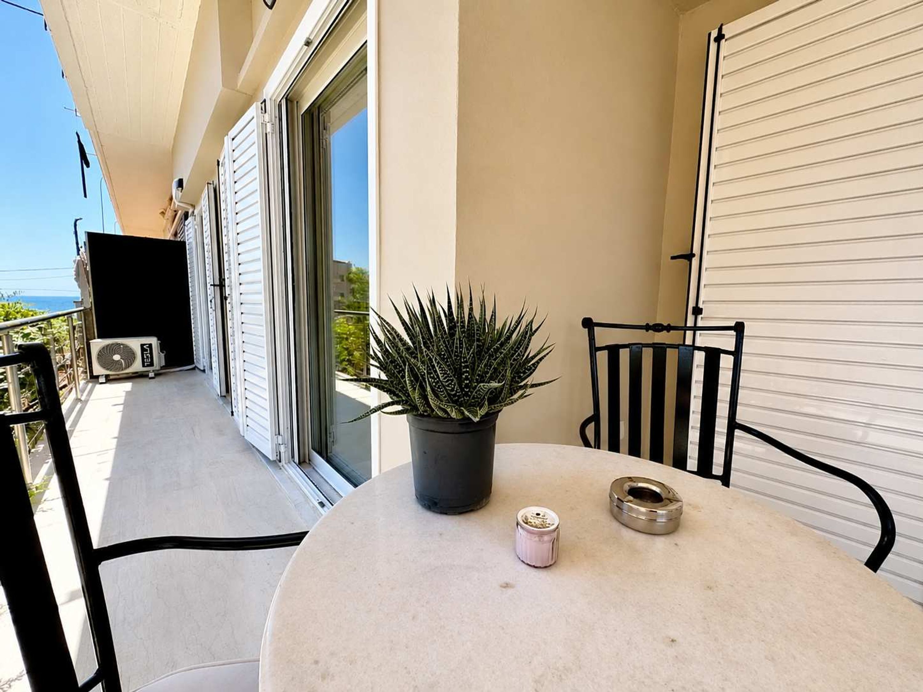 Un balcon avec un pot de fleurs, une table avec une bougie et un objet en métal, et un volet roulant fermé.