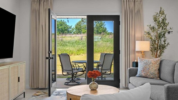 A modern room with a sofa, a dining table with chairs, a TV, and a view of a garden through a glass door. - Keystone Lake Getaway (Sand Springs)