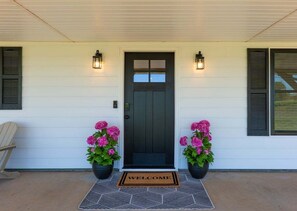 A white house with a dark front door, two potted pink flowers, and a welcome mat. - Keystone Lake Getaway (Sand Springs)
