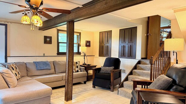 A living room with a sofa, armchairs, a coffee table, a lamp, a ceiling fan, and a staircase. - Scenic Wine Trail Lodge w/ Decks & Lake Views (Penn Yan)