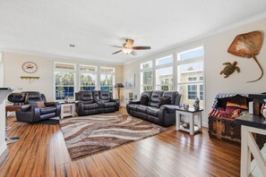 A spacious living room with leather sofas, a wooden floor, large windows, a ceiling fan, and decorative wall art. - The Blue Heron at River's Bend - Steinhatchee (Steinhatchee)