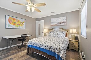Ein Schlafzimmer mit Bett, Schreibtisch, Stuhl, Nachttischen, Lampen, Deckenventilator und Wandbildern, darunter eine Karte und ein Landschaftsgemälde.