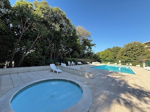 Ein Swimmingpool mit Liegestühlen und einer kleinen Whirlpoolanlage, umgeben von Bäumen und blauem Himmel.