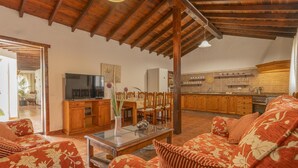 Sala de estar com televisão, mobiliário de madeira e cozinha ao fundo.