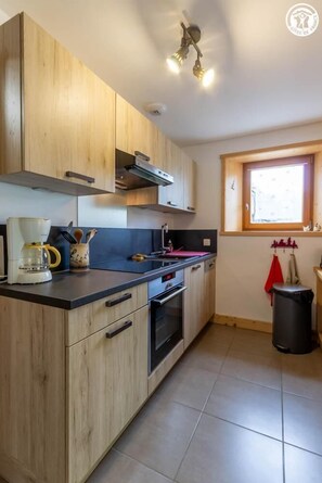 A kitchen with wooden cabinets, a coffee maker, a window, and a trash bin. - LA BARTAVELLE - LA GIETTAZ (LA GIETTAZ)