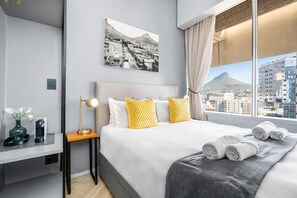 A hotel room with a bed, a desk, a lamp, a vase with flowers, and a cityscape view. - One Thibault 1609 - Studio Apartment, Sleeps 2 (Cape Town)