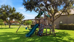A playground with a slide and swings, surrounded by grass and trees.