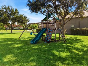 A playground with a slide and swings, surrounded by grass and trees.