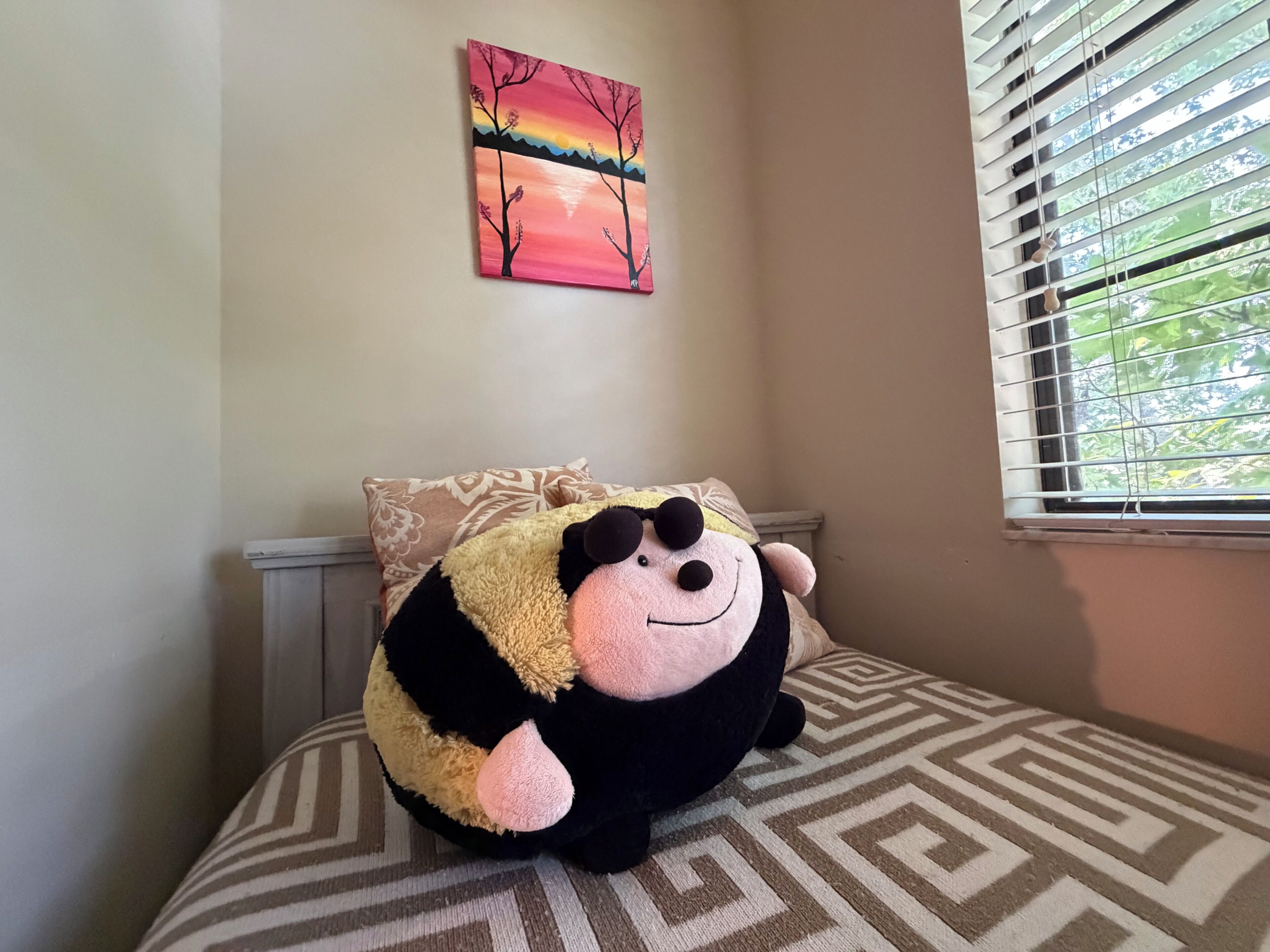 A bed with a patterned bedspread, a wall with a framed painting, and a window with blinds.