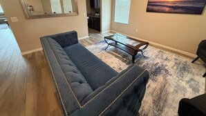A living room with a dark-colored sofa, a glass coffee table, a painting of a sunset, and a kitchen in the background.
