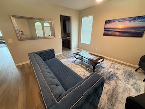 A living room with a dark-colored sofa, a glass coffee table, a painting of a sunset, and a kitchen in the background.