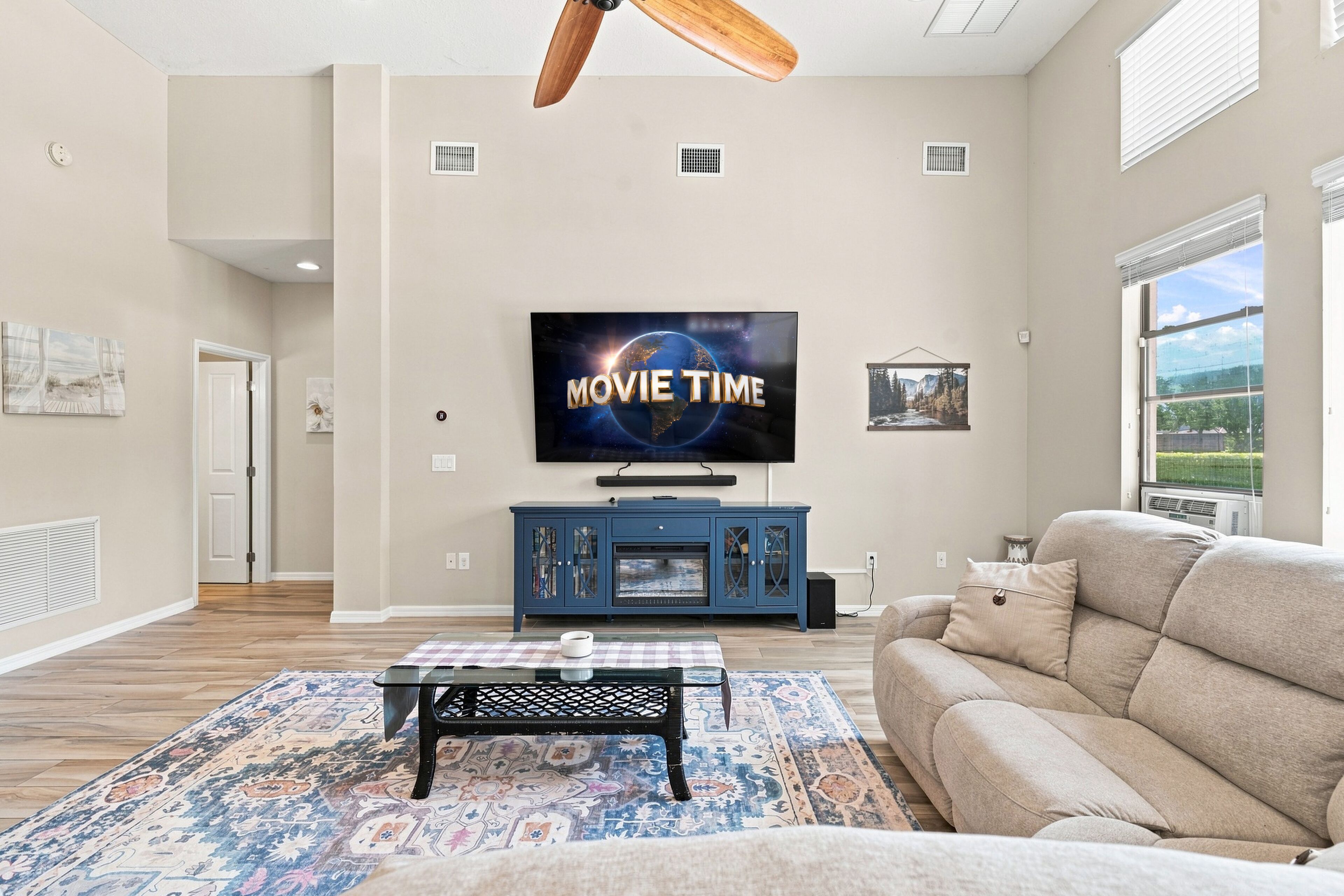 A living room with a flat-screen TV mounted on the wall, a blue entertainment unit, a beige sofa, a patterned rug, and a ceiling fan.