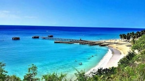 A beach with clear turquoise waters, a pier extending into the sea, and a sandy shore lined with palm trees.