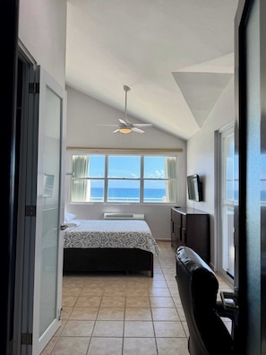 A bedroom with a bed, a ceiling fan, a window with curtains, and a view of the sea.