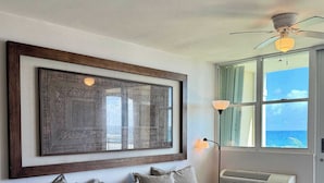 A living room with a grey sectional sofa, a large framed artwork, a ceiling fan, a window with a view, and an air conditioning unit.