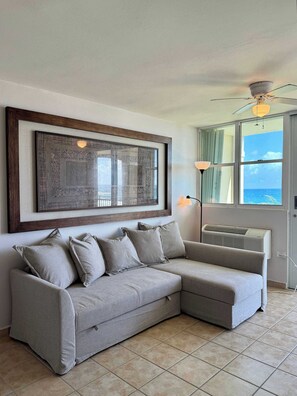 A living room with a grey sectional sofa, a large framed artwork, a ceiling fan, a window with a view, and an air conditioning unit.