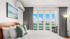 A hotel room with a bed, a painting, an air conditioner, a view of the city and water, and a lamp. - Sunlit Studio ° Private Balcony & Lagoon Views (San Juan)
