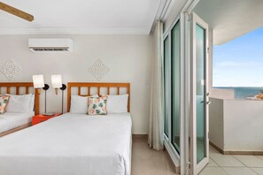 A hotel room with two beds, a ceiling fan, an air conditioning unit, decorative wall art, a sliding glass door to the balcony, and a view of the sea. - Breezy Studio ° Lagoon Views & Pool (San Juan)