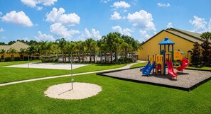 Ein Spielplatz mit Rutschen und einer Kletterkonstruktion, umgeben von einem gepflegten Rasen und einem gelben Gebäude im Hintergrund.
