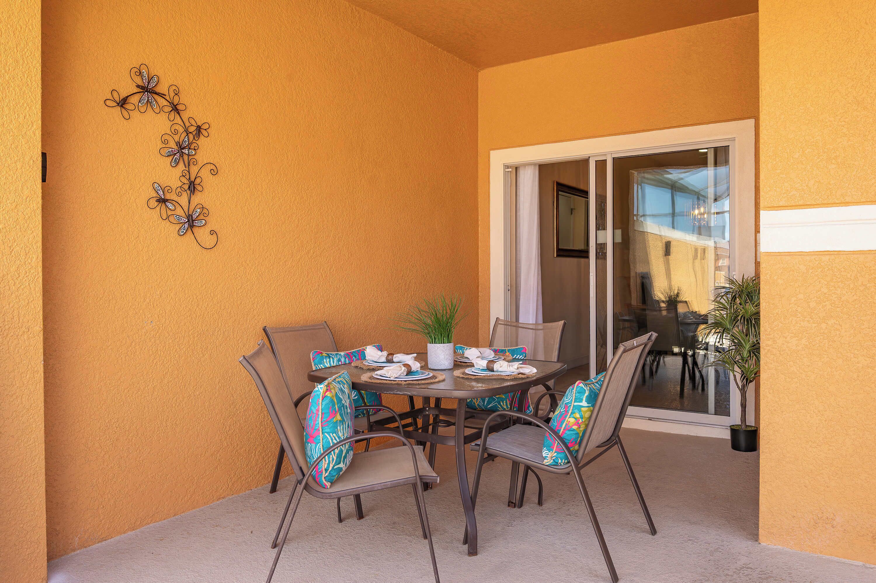 An outdoor dining area with a table and chairs, a decorative metal piece on the wall, and a sliding glass door leading to a patio.