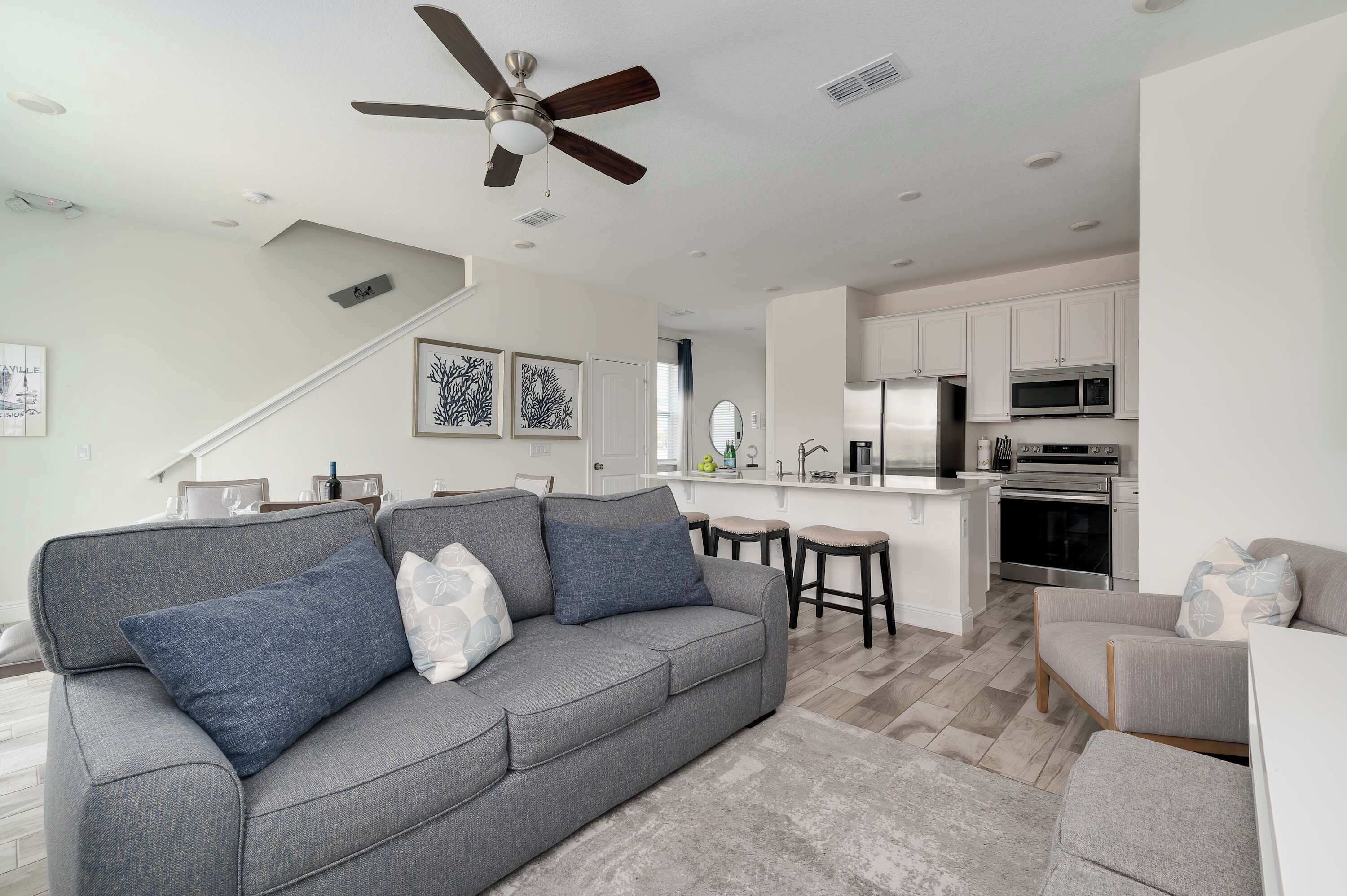 A modern living room with a grey sofa, a ceiling fan, and a kitchen area with white cabinets and appliances.