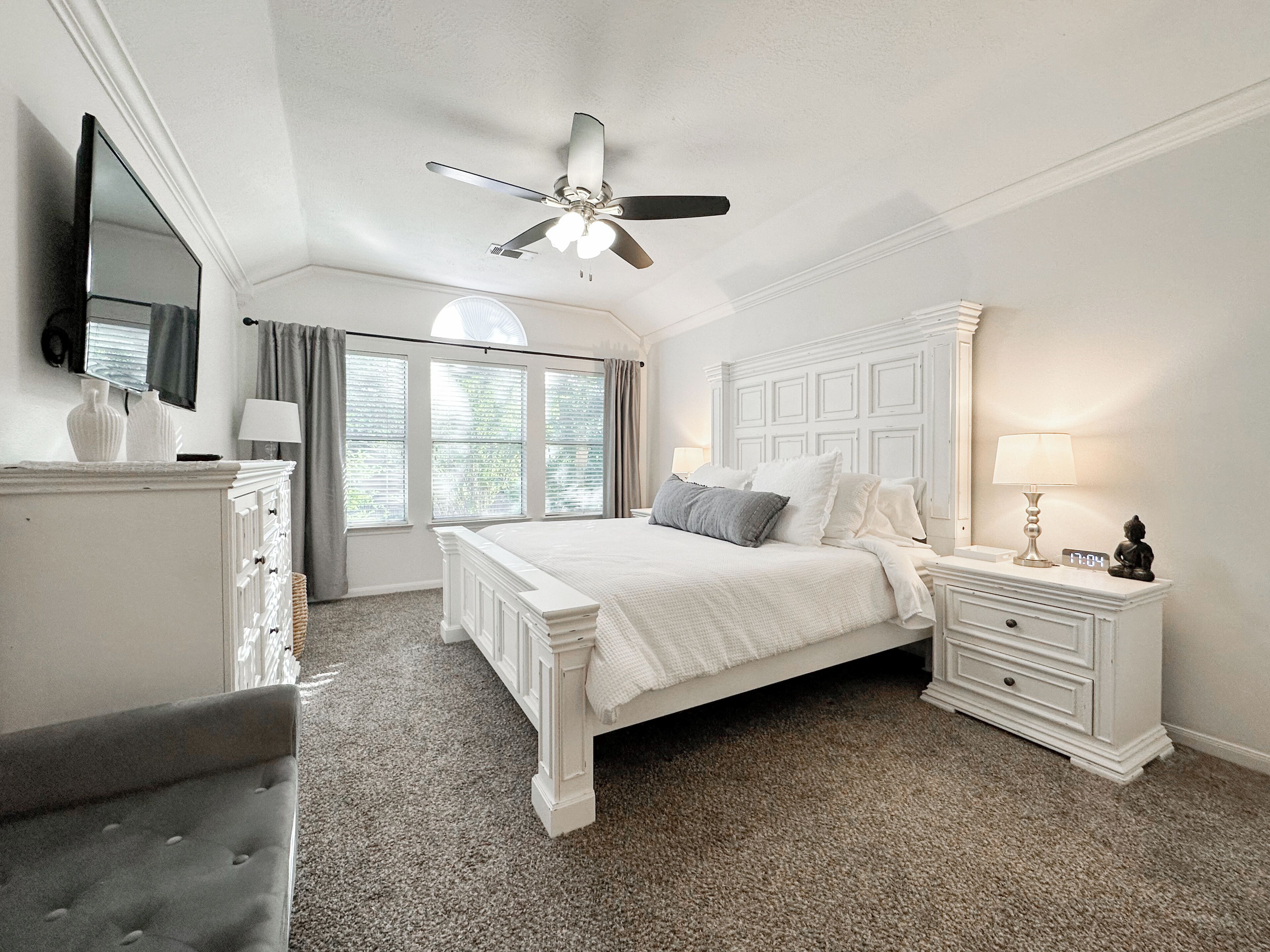 Une chambre à coucher comprenant un lit, une commode, une table de chevet, un ventilateur de plafond et une fenêtre avec des rideaux.