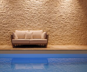Un canapé beige est placé contre un mur en pierre à la texture marquée, avec un bassin aux reflets scintillants en contrebas.