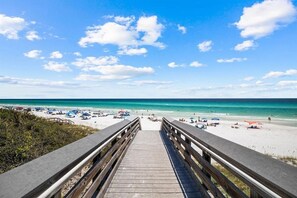 Ein hölzerner Steg führt zu einem Strand mit blauem Himmel und türkisblauem Wasser.