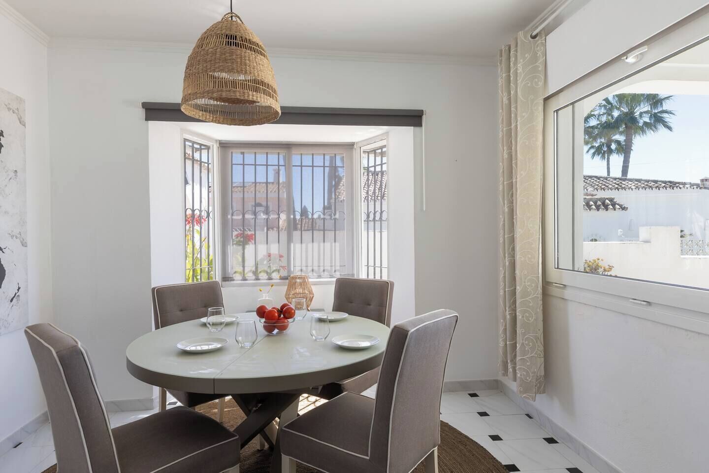 A dining area with a round table, four chairs, a hanging light fixture, and a view through a window.