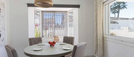A dining area with a round table, four chairs, a hanging light fixture, and a view through a window.