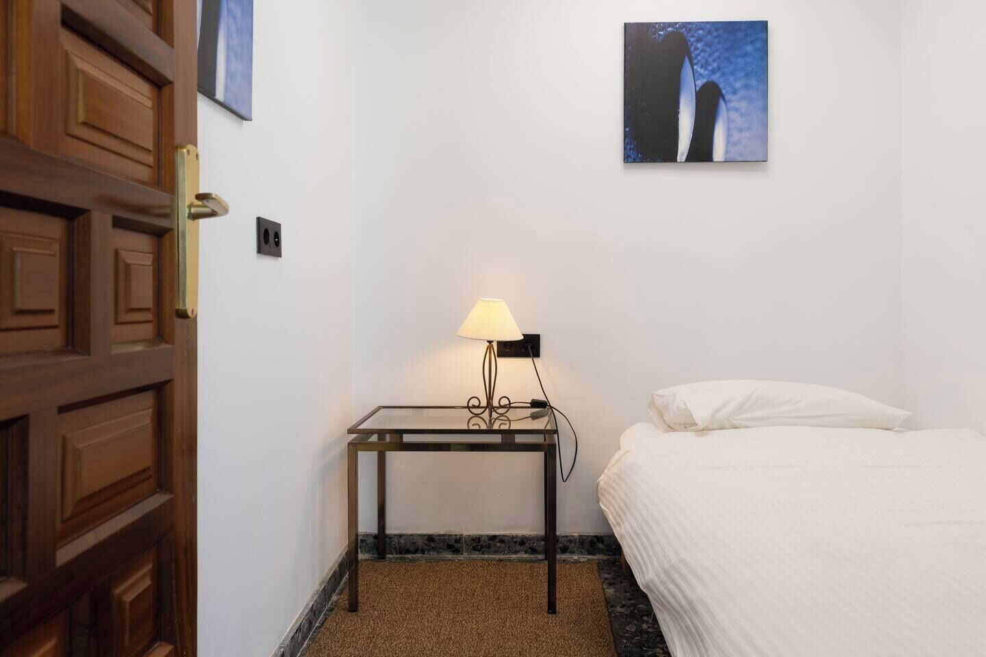 Et hotelværelse med en seng, et sengebord med lampe, et billede på væggen og en trædør.