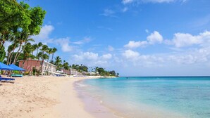 Ein Strand mit klarem türkisblauem Wasser, Palmen und einer Reihe von Strandgebäuden mit blauen Sonnenschirmen.