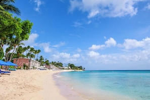 Ein Strand mit klarem türkisblauem Wasser, Palmen und einer Reihe von Strandgebäuden mit blauen Sonnenschirmen.